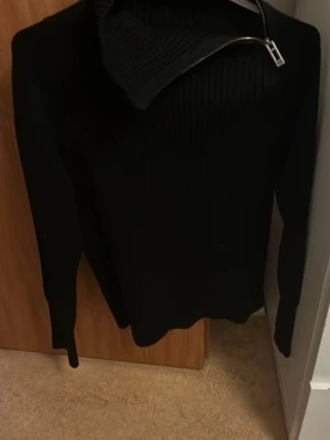 Svart stickad tröja med dragkedja - Säljer en svart stickad tröja med lång ärm och hög krage som har en snedställd dragkedja. Tröjan har en stilren och modern look, perfekt för dig som gillar minimalistiskt mode. Materialet ser mjukt och bekvämt ut, och modellen är något figurnära.