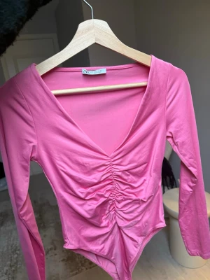 Rosa body med rynk framtill - Trendig rosa body från Zara med långa ärmar och djup V-ringning. Framsidan har snygg rynkning som ger en cool look. Tillverkad i mjukt stretchigt material som sitter skönt på kroppen. Perfekt att styla med jeans eller kjol. 
