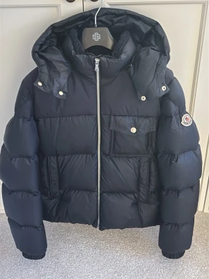 Svart dunjacka från Moncler - Snygg svart dunjacka från Moncler med huva och silverfärgad dragkedja. Jackan har en bröstficka med lock och knapp, samt Moncler-logga på ärmen. Puffig modell som håller dig varm under kalla dagar. 