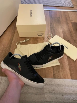 Svarta Axel Arigato sneakers med fågel - Snygga svarta sneakers från Axel Arigato i läder med broderad färgglad fågel på sidan. Klassisk låg modell med vita sulor och svarta skosnören. Perfekta för dig som vill ha en stilren men unik look. Storlek 41 passar 42 box medföljer ej