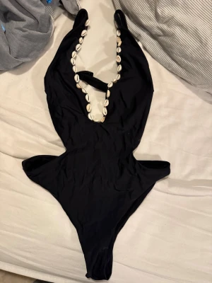 Svart baddräkt med snäckskal - Snygg svart baddräkt med djup urringning dekorerad med vita snäckskal längs banden. Modellen har cut-out detaljer i midjan och spänne i ryggen. Tillverkad i stretchigt material som passar perfekt för stranden eller poolen.