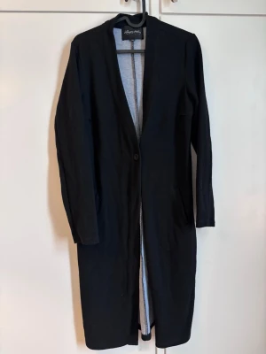 Svart lång kappa från Happy Holly - Stilren svart lång kappa från Happy Holly med enkel knäppning framtill och två fickor. Kappan har rak passform och är tillverkad i ett mjukt tyg som ger en clean look. Perfekt för dig som gillar minimalistisk stil. Strl 32/34