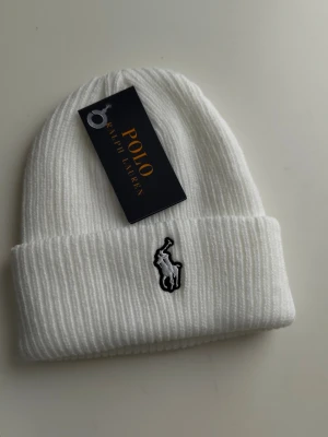 Vit ribbstickad mössa Polo Ralph Lauren - Snygg vit ribbstickad mössa från Polo Ralph Lauren med uppvikt kant och klassisk broderad logga framtill. Tillverkad i mjukt material som håller dig varm och stilren under kalla dagar. Perfekt accessoar för en clean och trendig look.