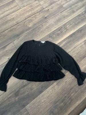 Svart volangblus från Gina Tricot - Snygg svart blus från Gina Tricot med diskreta prickar och flera volanglager framtill. Blusen har långa ärmar med knyt vid ärmsluten och rund halsringning. Perfekt för dig som gillar en trendig och lekfull stil.