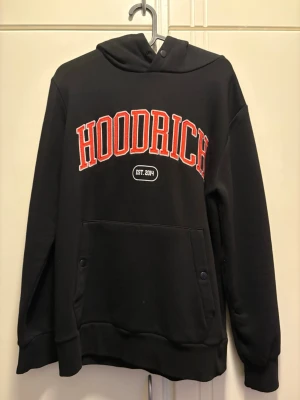 Svart hoodie från Hoodrich - Svart hoodie från Hoodrich med stor röd och vit logga på bröstet och textdetaljer på ryggen. Klassisk känguruficka framtill, ribbade muddar och huva. Tillverkad i mjukt bomullsmaterial, perfekt för en streetwear-look.