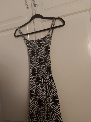 Svartvit långklänning med bladprint - Säljer en svart långklänning med vita bladmönster. Klänningen har smala axelband och en figurnära överdel som går ut i en vidare kjol. Perfekt för sommardagar och har en riktigt snygg och fräsch look.