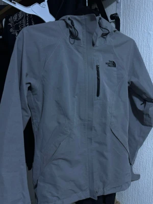 Grå vindjacka från The North Face - Snygg grå vindjacka från The North Face med huva och dragkedja framtill. Jackan har en bröstficka med dragkedja och två sidofickor. Tillverkad i slitstarkt material och har justerbar huva med snörning. Perfekt för blåsiga dagar.