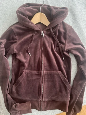 Brun velour hoodie från Juicy Couture - Mjuk och lyxig brun velour hoodie från Juicy Couture med dragkedja framtill, fickor och huva. Klassisk JC-logga broderad på bröstet och snygga metalldetaljer på snörena. Perfekt för en chill och trendig look.
