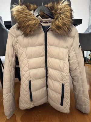 Beige pufferjacka med fuskpäls - Snygg beige pufferjacka med stor huva och fuskpälsdetalj. Jackan har svarta dragkedjor framtill och på fickorna, samt quiltade sömmar för extra stil. Perfekt för kalla vinterdagar och riktigt varm. Men den har ett skavmärke som visas på sista bilden.