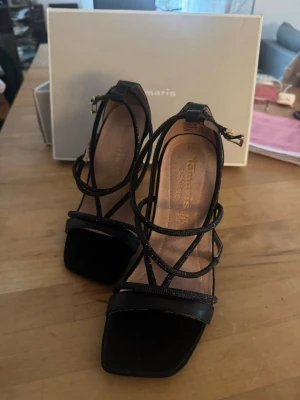 Svarta glittriga klackar från Tamaris - Snygga svarta pumps från Tamaris med smala, glittriga remmar och fyrkantig tå. Skorna har hög klack och justerbart spänne vid ankeln. Tillverkade i syntetmaterial med en elegant och modern design. Med en lila undersida 