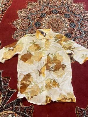 Blommig skjorta i beige och brunt - Säljer en luftig skjorta med blommigt mönster i beige, brunt och grönt. Skjortan har klassisk krage, knappar framtill och korta ärmar. Perfekt för dig som gillar en avslappnad och trendig stil.