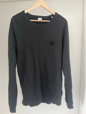 Svart sweatshirt från NNO7 - En stilren svart sweatshirt från NNO7 (No Nationality) med rund halsringning och diskret broderad logga på bröstet. Tröjan har långa ärmar och ribbade muddar vid ärmslut och nederkant. Perfekt för en avslappnad look.