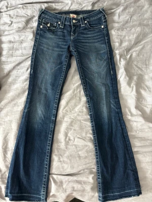 Blå bootcut jeans från True Religion - Snygga blå bootcut jeans från True Religion med klassiska kontrastsömmar och dekorativa fickor bak. Jeansen har låg midja, slitningar och detaljerade nitar. Perfekta för dig som gillar en avslappnad men trendig look.