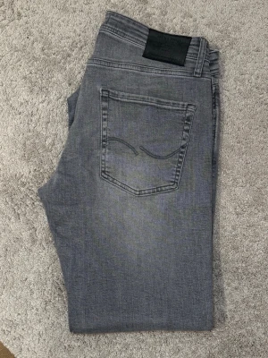 Grå slim fit tapered jeans från Jack & Jones - Snygga grå jeans från Jack & Jones i slim fit-modell. Jeansen har klassisk femficksdesign, diskret broderad bakficka och svarta detaljer vid linningen. Perfekta för en avslappnad och trendig look.