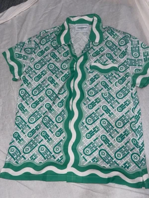 Grönt och vitt set från Casablanca - Snyggt set från Casablanca med skjorta och shorts i matchande grönt och vitt grafiskt mönster. Skjortan har korta ärmar, krage och knappstängning, medan shortsen har resårmidja med dragsko. Materialet ser ut att vara mjukt och glansigt, troligen polyester eller silkesliknande.