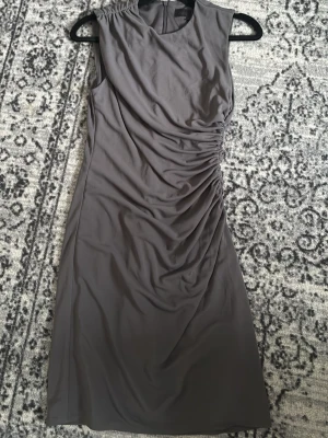 Grå omlottklänning från Zara - Snygg grå klänning från Zara med omlottdetaljer och rynkningar längs sidan. Ärmlös modell i mjukt, lätt material som ger en elegant siluett. Perfekt för dig som gillar stilrena och moderna plagg.