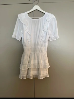 Vit volangklänning med kort ärm - Söt vit kortklänning med volangdetaljer och korta ärmar. Klänningen har v-ringning i ryggen, markerad midja och dubbla volanglager på kjolen. Tillverkad i ett lätt, luftigt tyg som passar perfekt till varma dagar. Klänningen är i strl 158/164