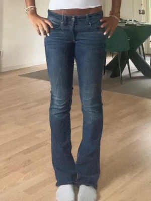 Blå bootcut jeans - Storlek 28/34 Säljer ett par blå jeans med bootcut passform och låg midja. Jeansen har klassisk femficksdesign, snygga slitningar och är tillverkade i ett mjukt jeansmaterial. Perfekta för dig som gillar en tidlös och trendig look.