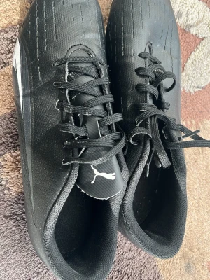 Svarta Puma sneakers med snörning - Säljer ett par svarta sneakers från Puma med klassisk snörning och diskret logga på plösen. Skorna har en låg profil och ovandel i textilmaterial med subtila sömdetaljer. Perfekta för dig som gillar stilrena och sportiga skor.