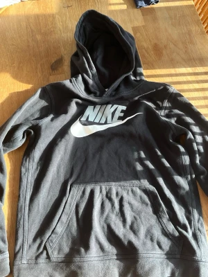 Svart Nike hoodie med stor logga - Säljer en svart hoodie från Nike med stor vit logga och swoosh på bröstet. Hoodien har en klassisk känguruficka framtill och huva. Tillverkad i mjukt bomullsmaterial som är skönt att ha på sig. Perfekt för dig som gillar sportig och avslappnad stil. Använt väldigt få gånger väldigt bra skick.