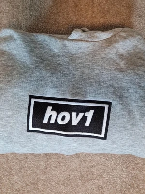 Grå hoodie med hov1-tryck - Snygg grå hoodie med stort hov1-tryck på bröstet i svart och vitt. Klassisk passform med huva och mjukt material som känns skönt mot huden. Perfekt för dig som gillar streetwear och vill ha en enkel men stilren look.