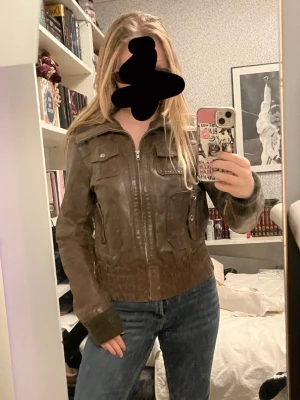 Brun skinnjacka med fickor - Snygg brun skinnjacka med dragkedja framtill och två bröstfickor med lock. Jackan har ribbade muddar vid ärmslut och midja för en tajtare passform. Perfekt till jeans och ger en cool look.