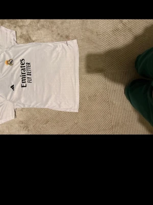 Vit Real Madrid fotbollströja - Säljer en vit Real Madrid fotbollströja från Adidas med klubbmärke och sponsortryck på bröstet. Tröjan har korta ärmar och är tillverkad i ett lätt och ventilerande polyestermaterial. Nummer 7 och spelarnamn på ryggen.