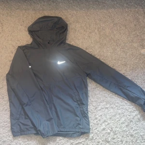 Nike Miler Repel Set - Snygg grå vindjacka från Nike med huva och hel dragkedja framtill. Jackan har långärmade ärmar, två sidofickor och en vit Nike-logga på bröstet. Perfekt för träning och chill, lätt och smidig att bära. Modern passform och sportig look. OBS! Byxorna är XL men passar L!