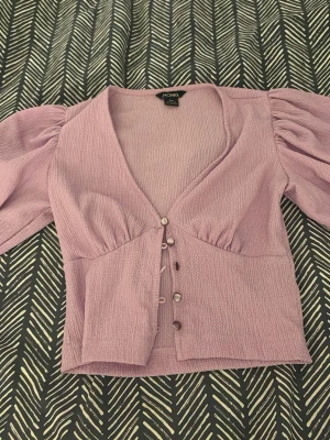 Ljuslila blus med puffärm från Monki - Supersöt ljuslila blus från Monki med puffiga ärmar, v-ringning och knappar framtill. Tillverkad i ett strukturerat tyg som ger en cool känsla. Perfekt att styla med jeans eller kjol för en trendig look.
