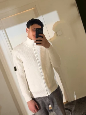 Vit stickad ziptröja från Stone Island - Vit stickad ziptröja från Stone Island med ribbad struktur och hög krage. Tröjan har dragkedja framtill och den klassiska svarta logotypen på vänster ärm. Perfekt för dig som gillar stilrena och exklusiva plagg.