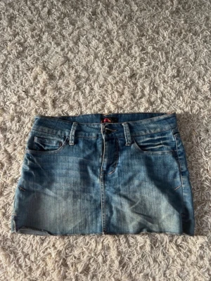 Blå lågmidjad jeanskjol med fickor - Säljer en klassisk blå jeanskjol i kort modell med fem fickor och bälteshällor. Kjolen har en snygg tvättad look och stängs med knapp och dragkedja framtill. Perfekt att matcha med en t-shirt eller hoodie.