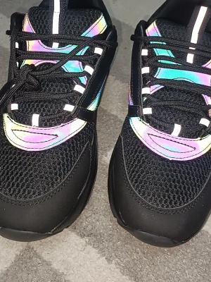 Svarta sneakers med reflekterande detaljer - Säljer ett par svarta sneakers med mesh-ovandel och färgglada, reflekterande detaljer i regnbågstoner. Skorna har svarta snören och en chunky sula som ger en modern vibe. Perfekta för dig som vill sticka ut med coola effekter när ljuset träffar skorna. Använda en gång i några timmar💫