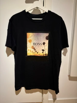 Svart BOSS t-shirt med palmtryck - Snygg svart t-shirt från BOSS med färgglatt palmtryck och logga på bröstet. Klassisk rund hals och korta ärmar. Perfekt för dig som gillar stilrena plagg med en twist. Tillverkad i mjuk bomull för skön känsla.