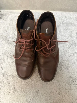 Bruna läderloafers från Timberland - Säljer ett par klassiska bruna loafers från Timberland i äkta läder. Skorna har rundad tå, snörning framtill och en stilren design som passar till många olika outfits. Mjukt innerfoder och robust yttersula för extra komfort.