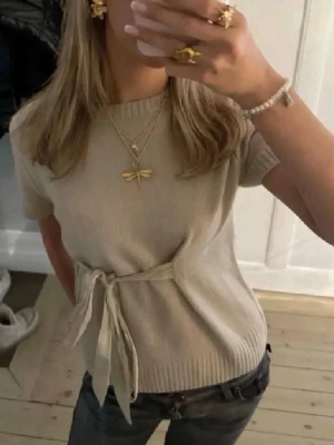 Beige stickad topp med knyt från Zara - Superfin beige stickad topp från Zara med korta ärmar och rund halsringning. Toppen har en snygg knyt-detalj i midjan. Använd en gång!