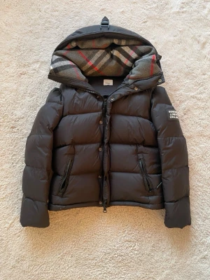 Svart pufferjacka från Burberry - Säljer en svart pufferjacka från Burberry med avtagbara ärmar och huva. Jackan har klassiskt rutigt foder i huvan, två dragkedjefickor och Burberry-logga på ärmen. Tillverkad i glansigt material och perfekt för kalla vinterdagar.
