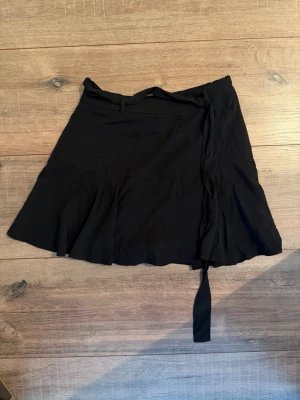 Svart kjol (shorts under) - Säljer en svart kort kjol med knytband i midjan. Kjolen har en lätt utsvängd form och är perfekt för dig som gillar en enkel men snygg stil. Materialet känns mjukt och följsamt, troligen polyester eller bomull. Passar till många olika looks!