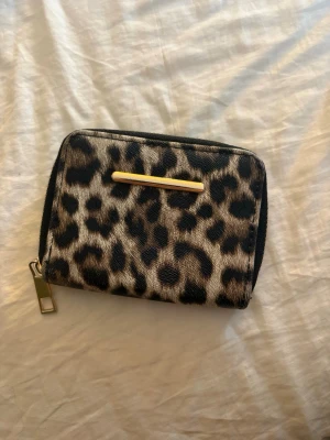 Leopardmönstrad plånbok från Primark - Snygg plånbok från Primark med leopardmönster i beige, brunt och svart. Plånboken har guldfärgad dragkedja och dekorativt metallband på framsidan. Insidan har flera fack för kort och sedlar. Materialet är konstläder med textilinsida.