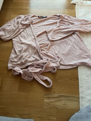 Ljusrosa morgonrock med knytband - Mysig ljusrosa morgonrock i mjukt tyg med långa ärmar och tillhörande knytband i midjan. Perfekt för att chilla hemma och svepa in sig i efter duschen. Enkel och stilren design utan mönster.