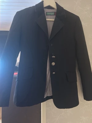 Svart kavaj från Equipage - Stilren svart tävlingskavaj till barn från Equipage med klassisk passform och tre dekorativa silverknappar framtill. Kavajen har två fickor med lock och är helfodrad. En knapp saknas (översta) och en knapp hänger löst men går att sy fast igen