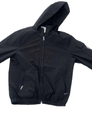 Svart vindjacka från Moncler med huva - Snygg svart vindjacka från Moncler med dragkedja framtill, två fickor med dragkedja och elastisk nederkant. Jackan har huva och en liten ficka på ena ärmen. Perfekt för blåsiga dagar och stilren look.