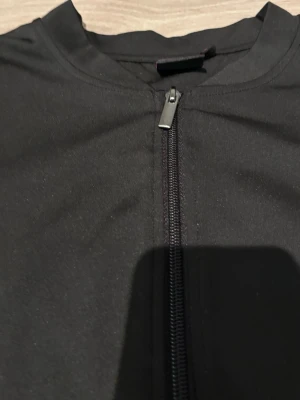 Svart träningsjacka från Phelix Sports - Snygg svart träningsjacka från Phelix Sports med hel dragkedja framtill. Tillverkad i ett stretchigt och funktionellt material som passar perfekt till träning eller uppvärmning. Enkel design med diskret logga i nacken.