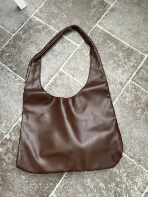 Brun axelväska i skinnimitation - Stilren brun axelväska i skinnimitation med enkel design och bred axelrem. Väskan har en mjuk, slät yta och rymligt huvudfack. Perfekt för dig som vill ha en minimalistisk och trendig accessoar till vardagslooken.Helt nyDjur och rökfritt hem 