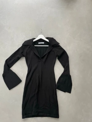 Svart stickad klänning från Adore - Svart stickad klänning från Adore med v-ringning, bred krage och långa trumpetärmar. Klänningen är figurnära och har en kort längd som ger en trendig och cool look. Perfekt för dig som gillar stilrena och moderna plagg.