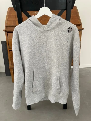 Grå hoodie med broderade detaljer - Snygg grå hoodie med huva och känguruficka framtill. På bröstet finns broderade stjärnor och en liten broderad fjäril som ger en unik touch. Tillverkad i mjukt sweatshirtmaterial som är perfekt för chill dagar. Passar dig som gillar enkel men ändå personlig stil.