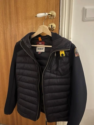 Svart dunväst från Parajumpers - Snygg svart dunväst från Parajumpers med quiltad framsida, gul detalj vid dragkedjan och klassisk logga på axeln. Västen har en innerficka med orange kant och är tillverkad i ett slitstarkt material med dunfyllning för extra värme.