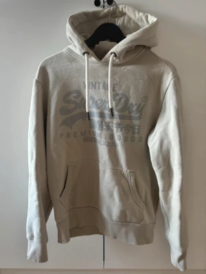 Beige hoodie från Superdry - Snygg beige hoodie från Superdry med stor logga och japansk text på bröstet. Klassisk känguruficka framtill, ribbade muddar och justerbar huva med vita snören. Tillverkad i mjukt bomullsmaterial som känns skönt mot huden.