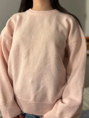 Ljusrosa stickad tröja - Mysig ljusrosa stickad tröja med rund halsringning och breda ribbade muddar vid ärmslut och nederkant. Tröjan har en avslappnad passform och är perfekt för lager på lager. Passar till jeans eller kjol för en enkel och stilren look.