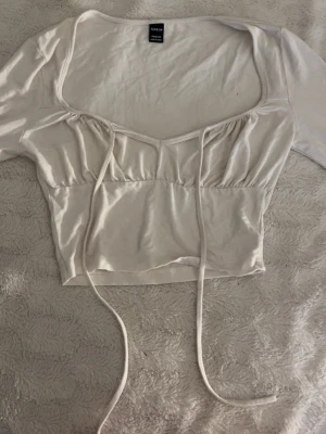 Vit croppad topp med knyt från SHEIN - Trendig vit croppad topp från SHEIN med långa ärmar och snygg knytning framtill. Toppen har en djup v-ringning och är tillverkad i ett mjukt material som känns skönt mot huden. Perfekt för att skapa en cool och avslappnad look.