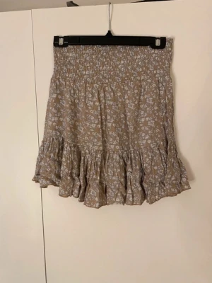 Beige blommig volangkjol - Supersöt beige kjol med vitt blommönster och bred smock i midjan. Kjolen har volangkant nedtill som ger en flowy och avslappnad look. Perfekt för sommardagar och enkel att matcha med en enkel topp. Materialet känns lätt och luftigt. Använd endast en gång och är fin i kvaliten.